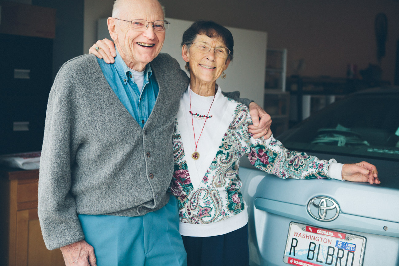 A husband and wife in their 80s laugh and hug as they stand next to their car