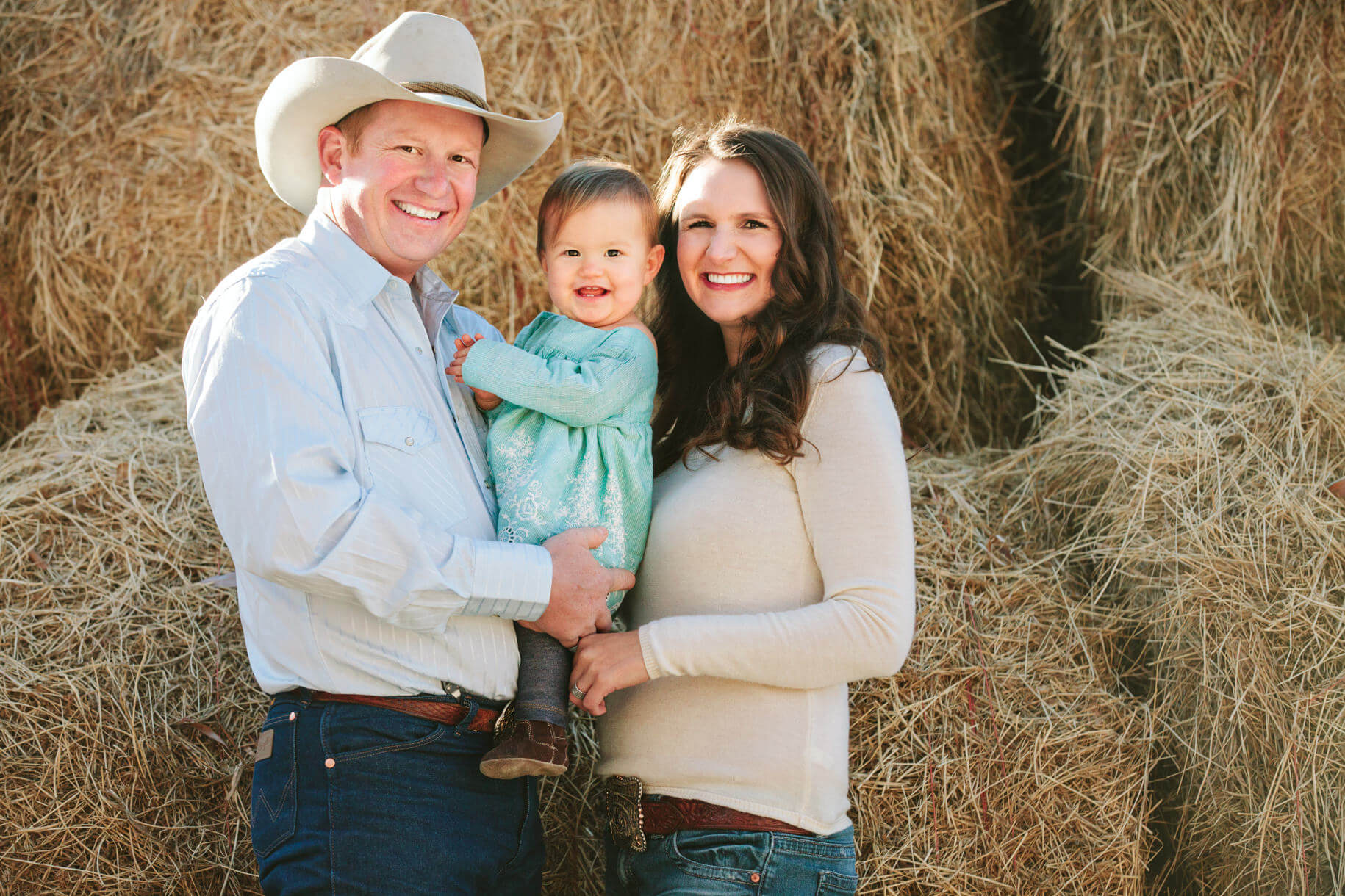 A mother and father hold their toddler daughter in front of hay bales during their family photos in Stevensville Montana