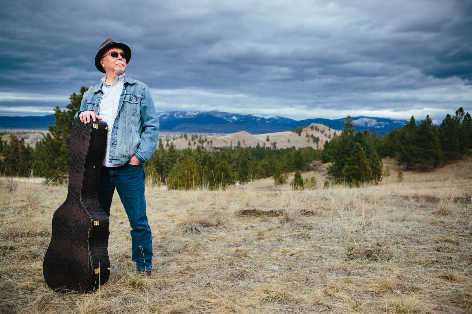 A musician stands in a field with his guitar case during his Missoula Montana portrait session