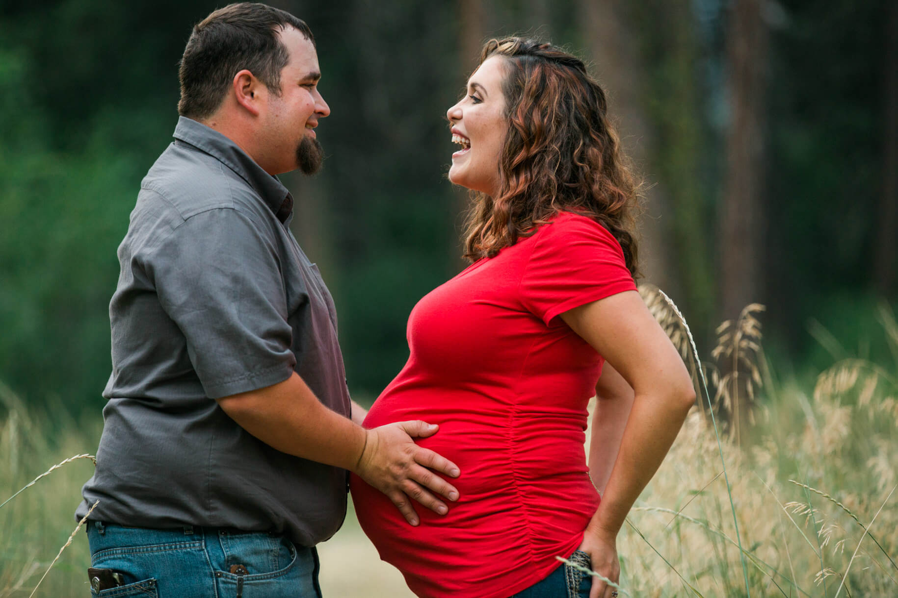 A father holds his pregnant wife's belly and laughs during their maternity photo session in Missoula Montana