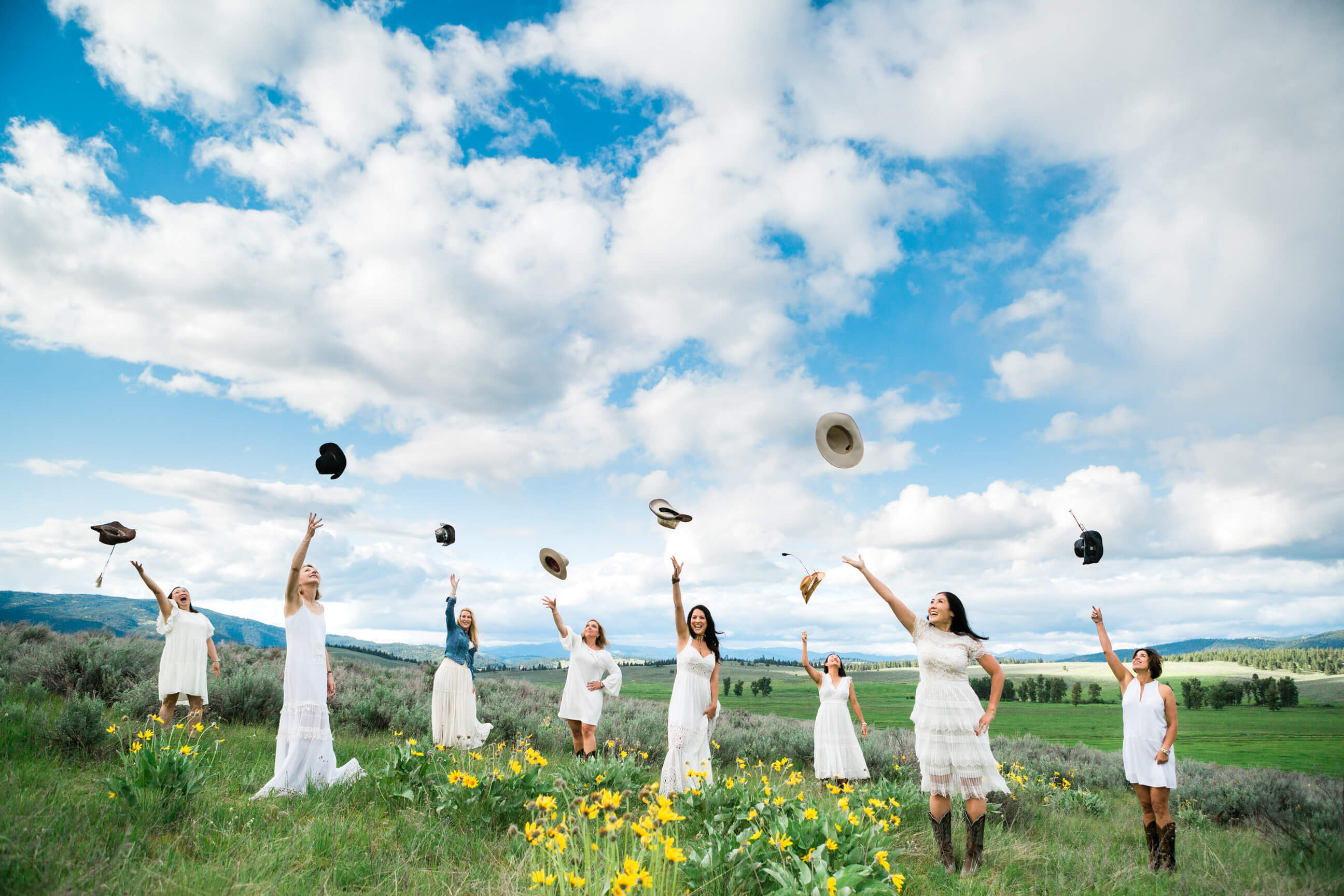 A group of women friends wearing white dresses stand in a field of wildflowers and toss their hats into the air at Paws Up Resort in Montana