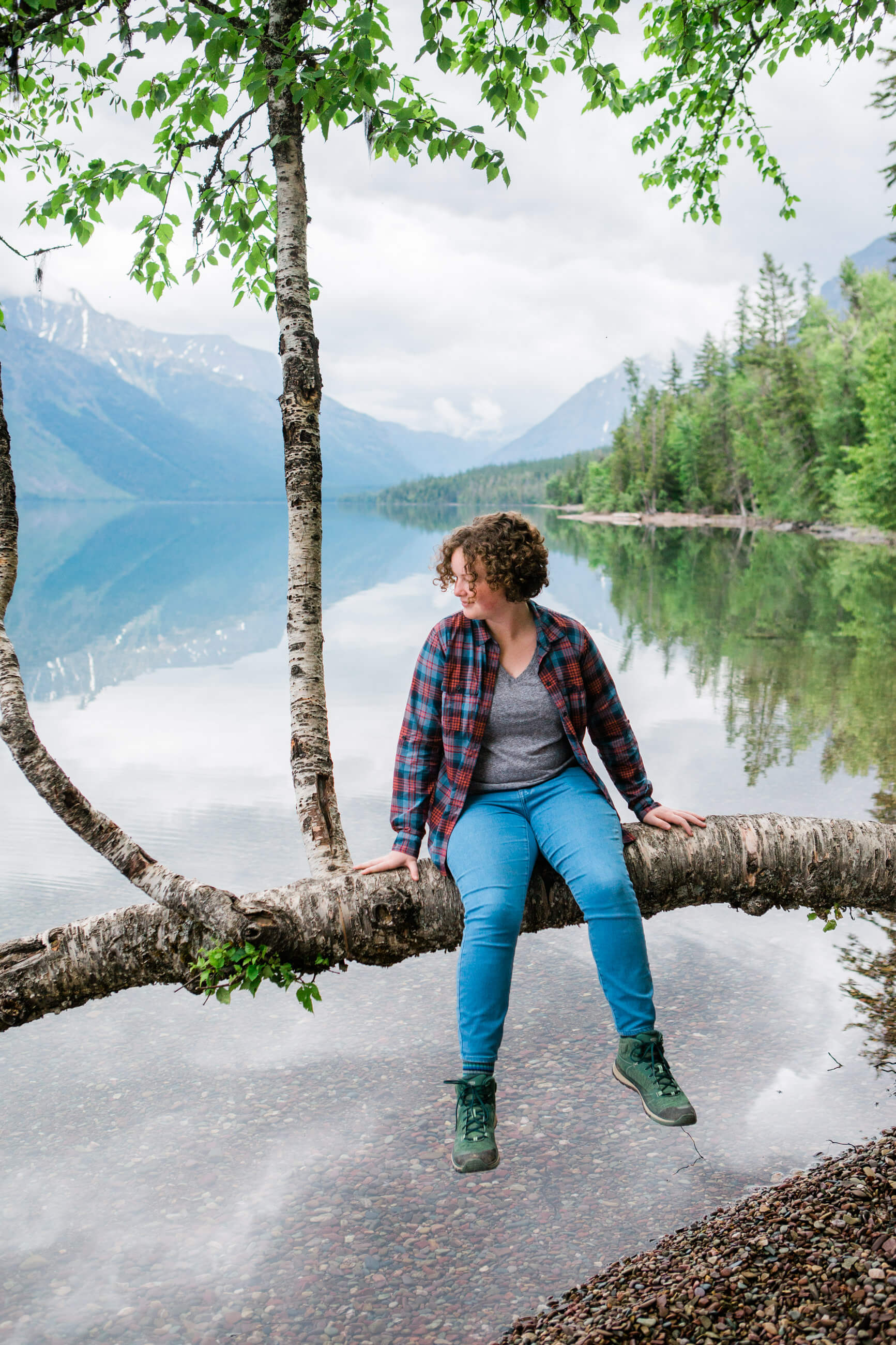 A teenage girl sits on a tree over Lake McDonald in Glacier National Park in Montana during her family photo session