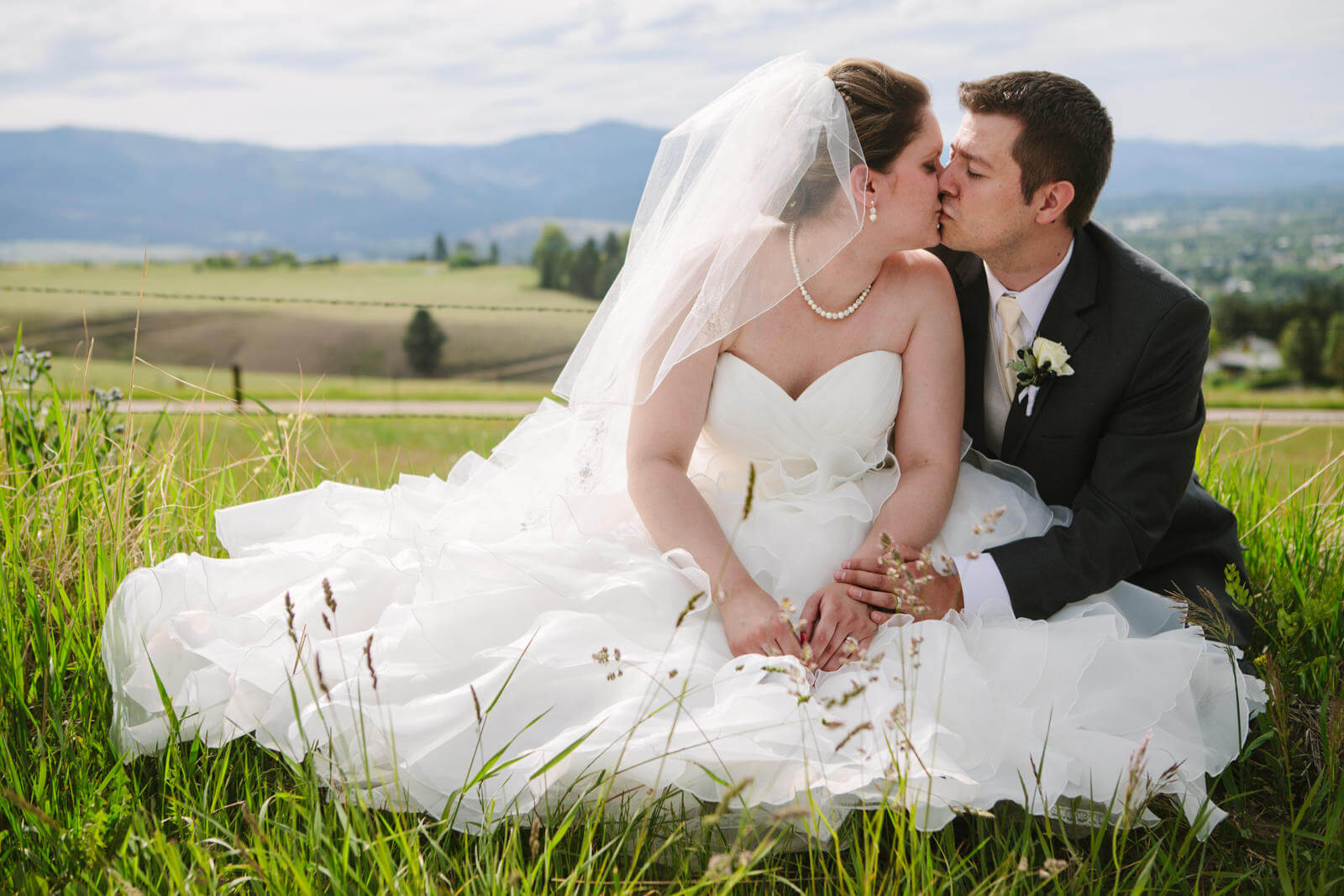 A bride and groom sit in grass and kiss during their wedding in Missoula Montana.