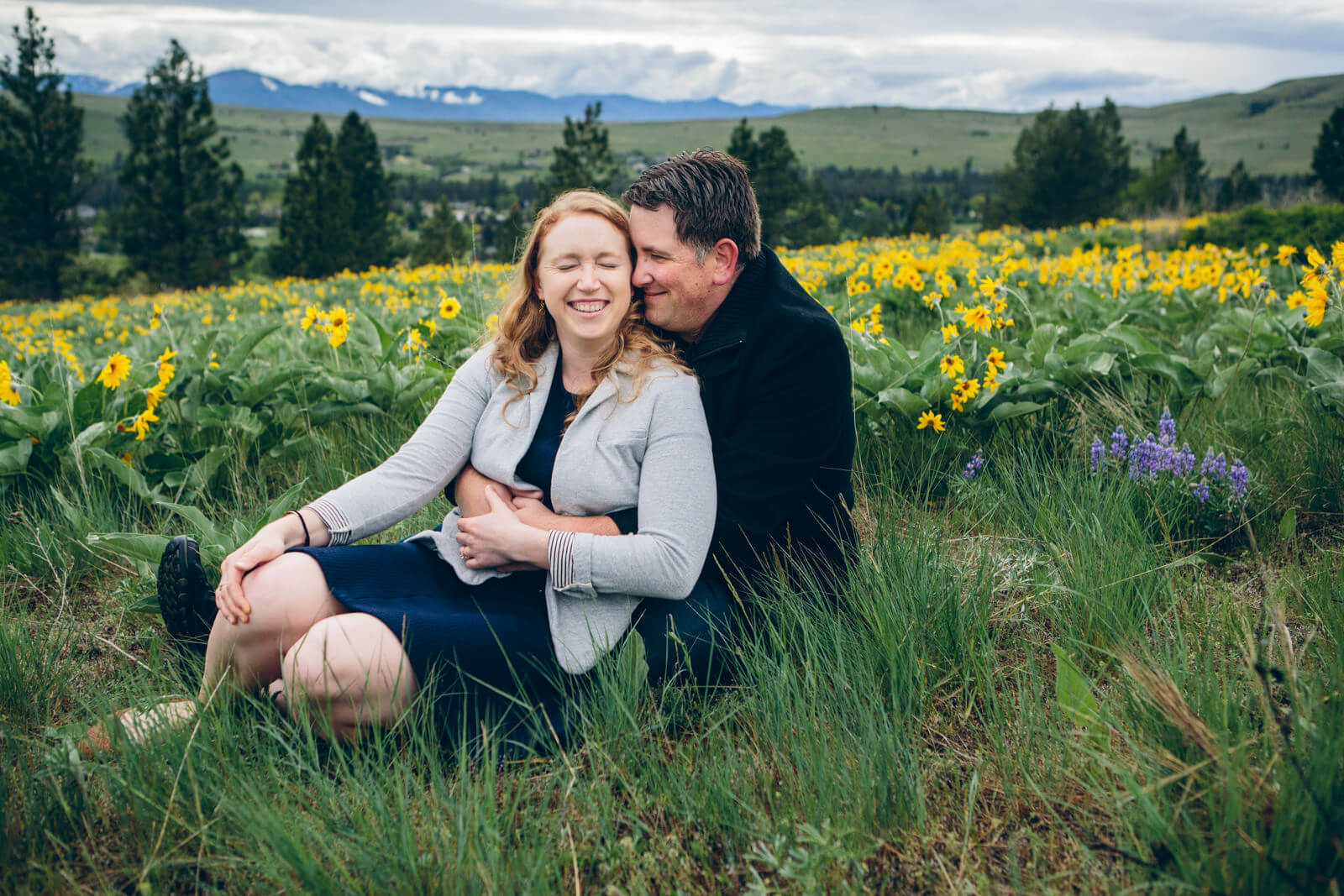 An engaged couple embraces and laughs amidst a field of arrow leaf balsam root wildflowers during their spring engagement session in Missoula Montana