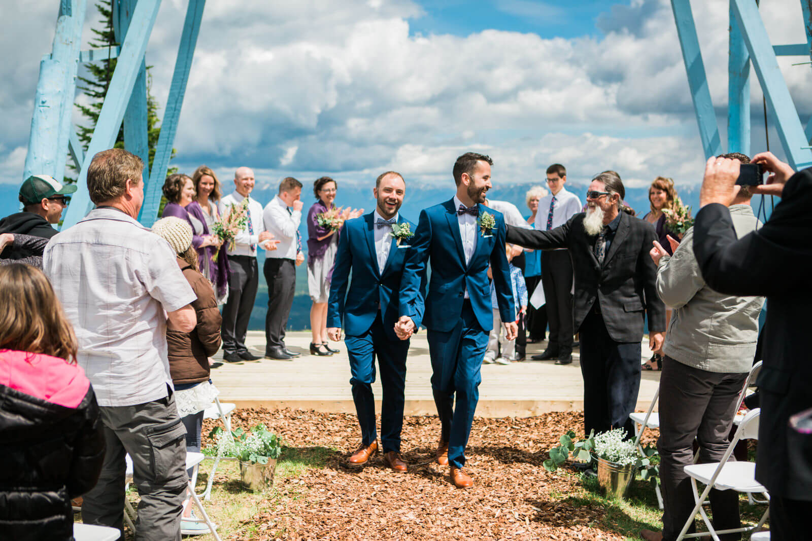 Two grooms walk down the aisle holding hands and smiling at their wedding in Kimberley, British Columbia, Canada.