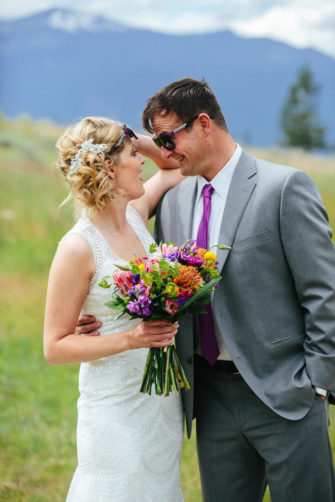 A bride and groom wearing sunglasses laugh with one another in front of a mountain backdrop at their wedding at Stone Tower Estates in Stevensville Montana