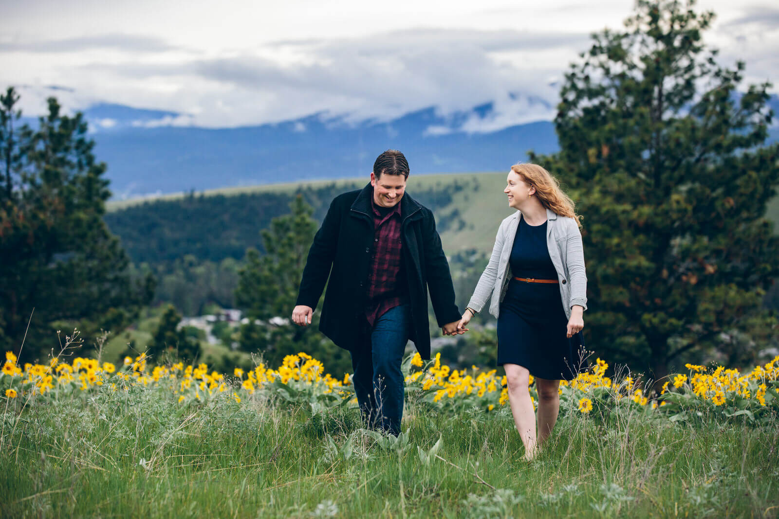 A couple walks through arrow leaf balsam root wildflowers during their engagement session in Missoula Montana