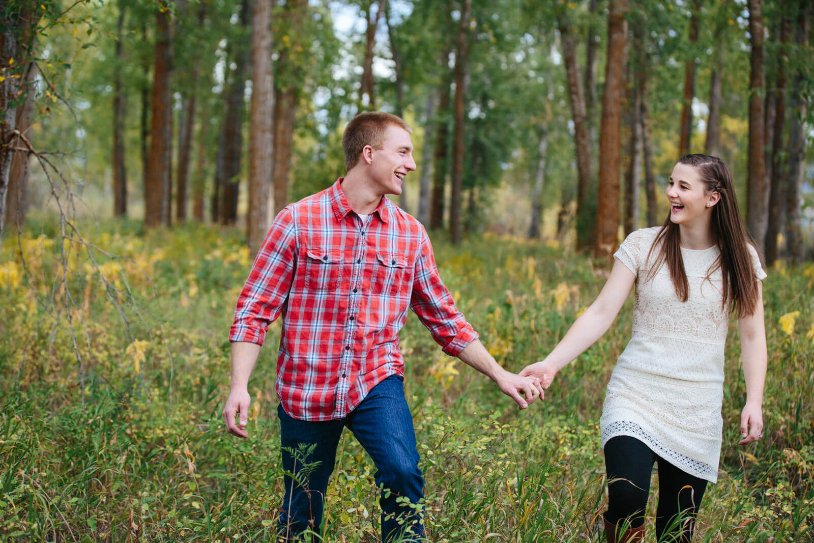A couple holds hands and laughs as they walk through the woods during their engagement session in Missoula Montana