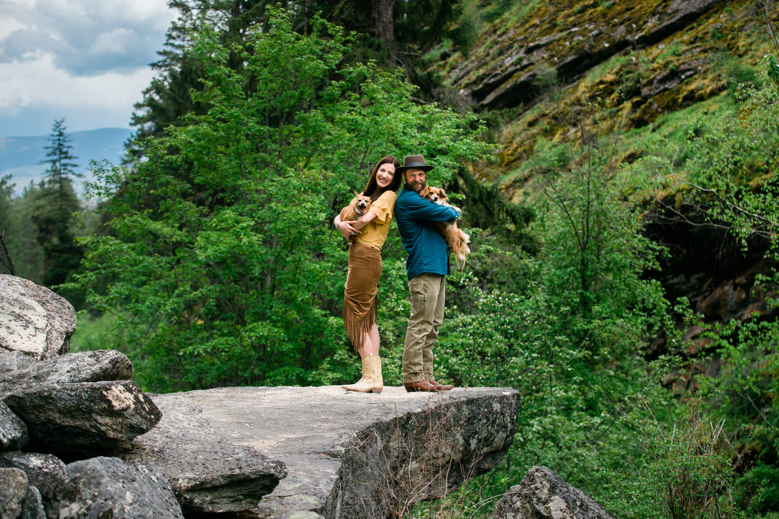 A couple holds their dogs while standing on a rocky outcrop during their engagement photos in the Bitterroot Valley in Montana