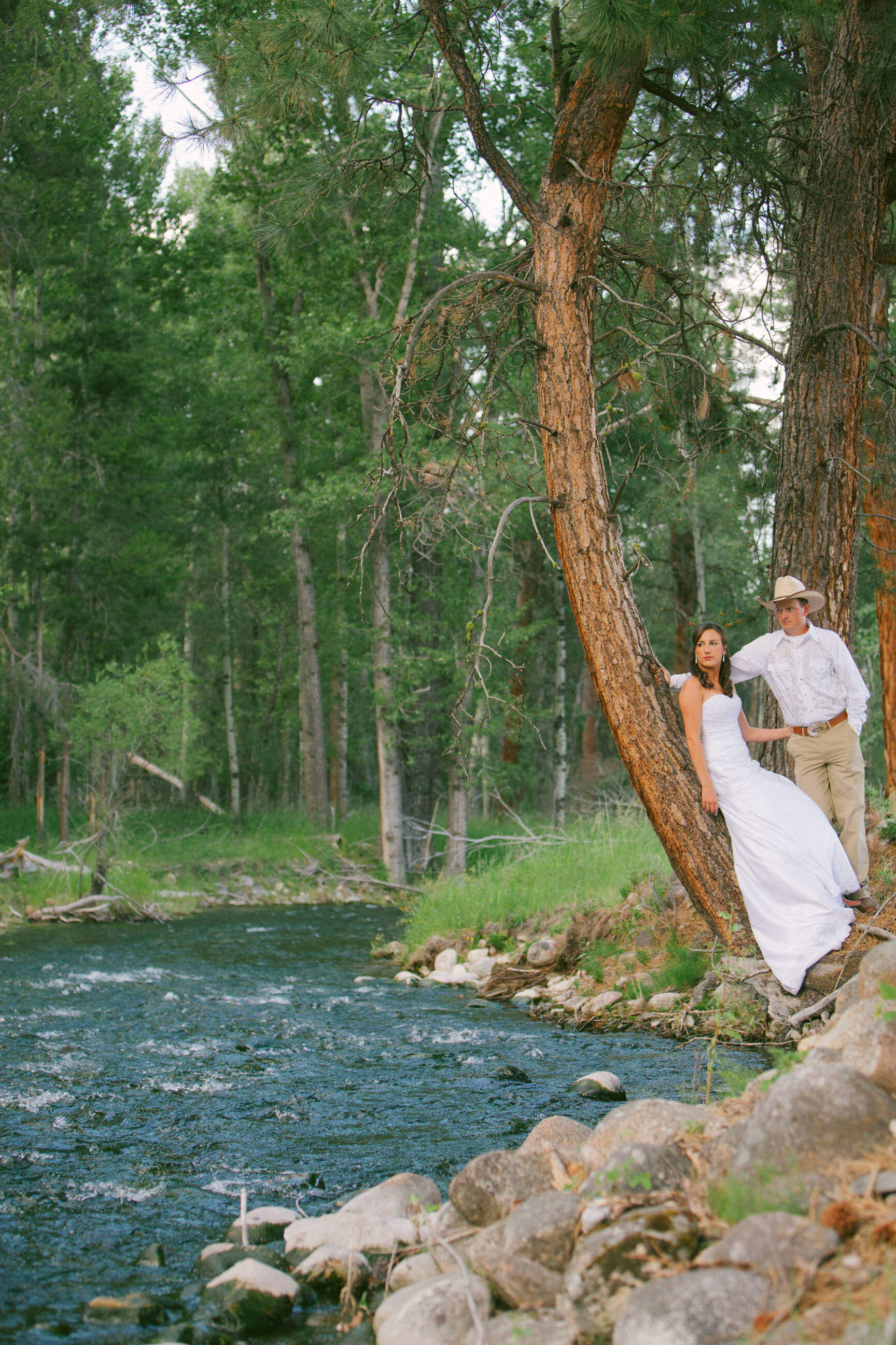 A bride and groom lean on a tree near a creek during their rock the dress photos on their ranch in Darby Montana