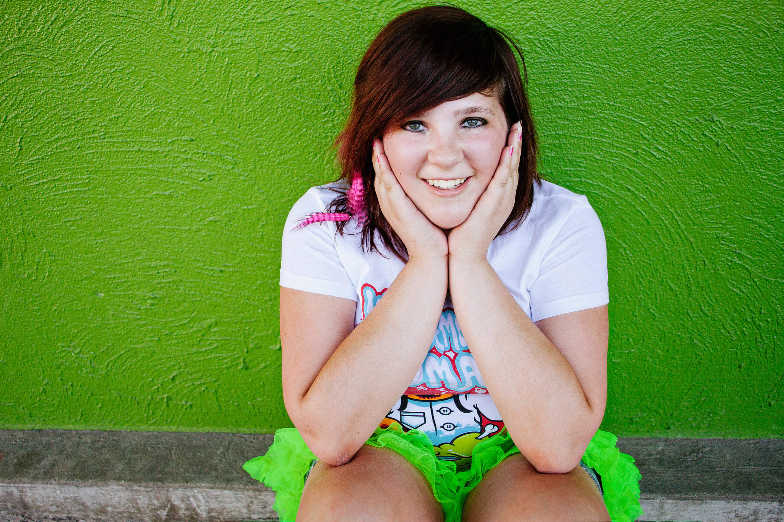 A senior girl wearing a neon green tutu smiles and sits near a green wall at her senior portraits in Missoula Montana
