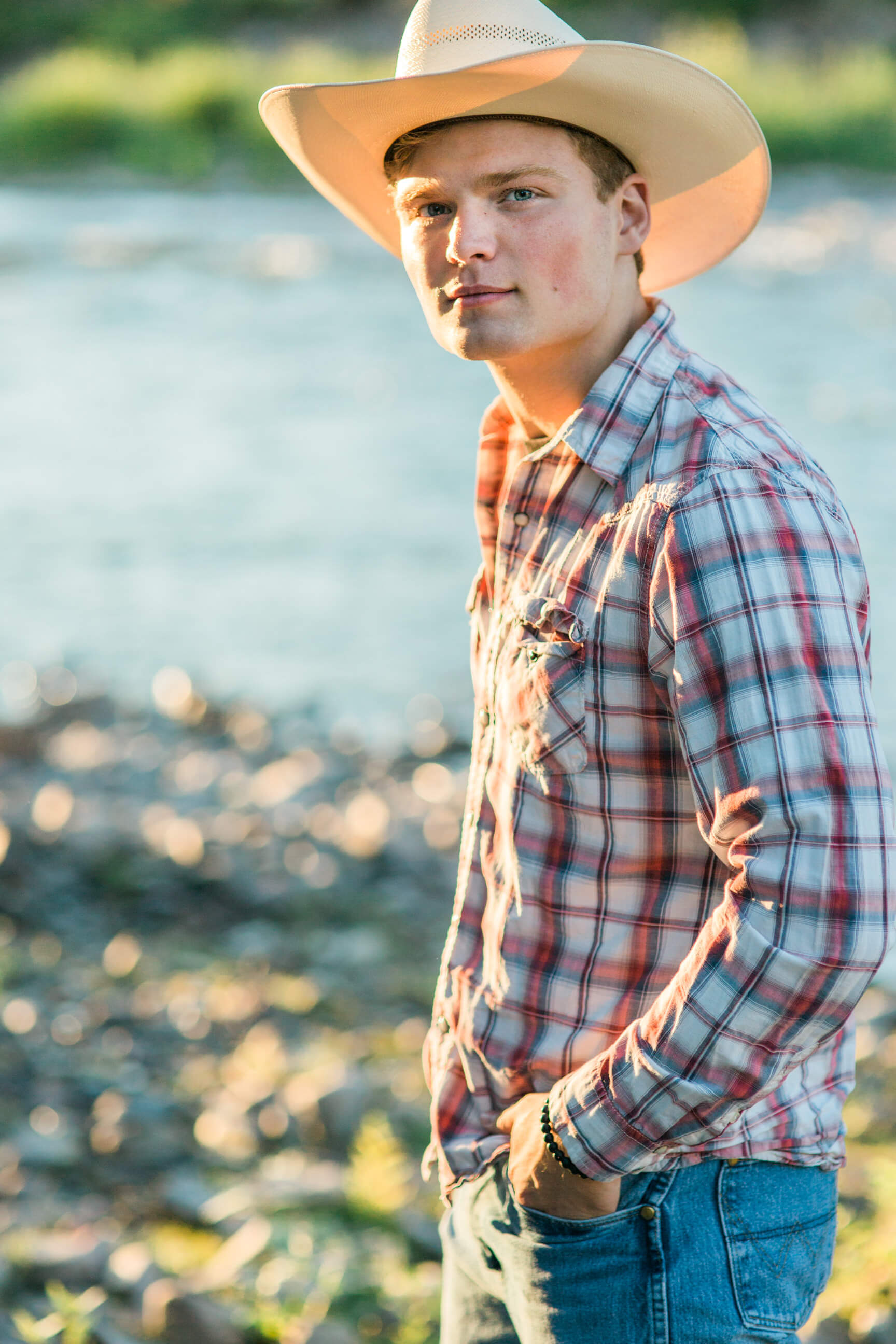 A senior boy wearing a cowboy hat looks at the camera as he stands near the Clark Fork River during his senior photos in Missoula Montana