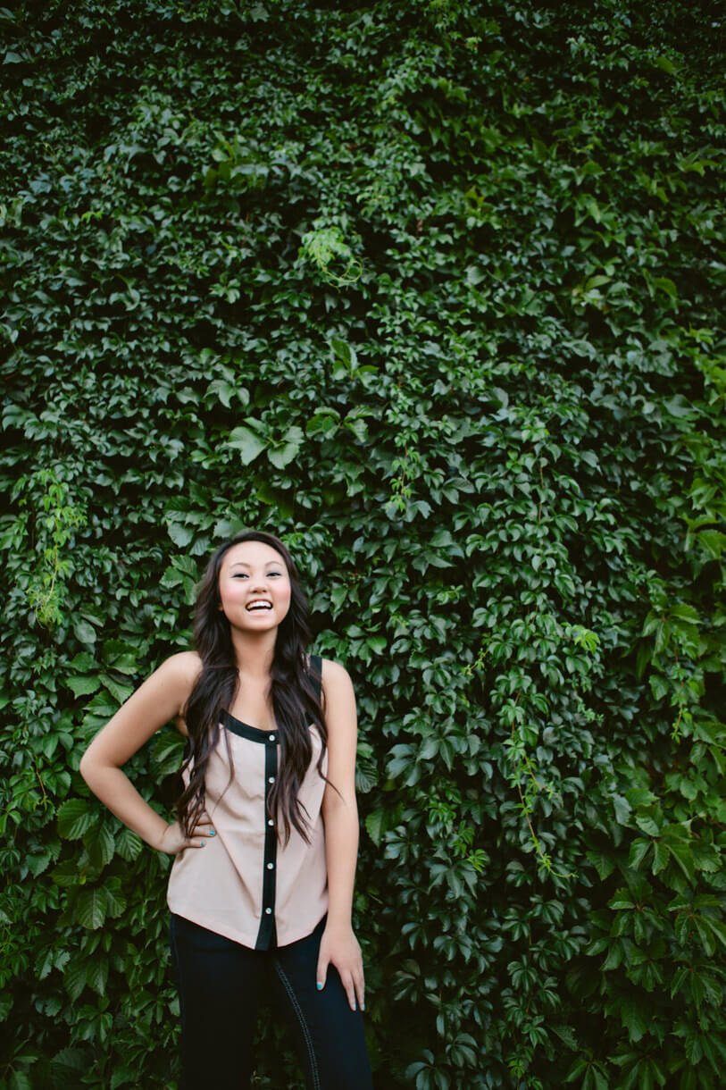 A high school senior girl smiles and laughs against a wall of green leaves during her senior photos in Missoula Montana