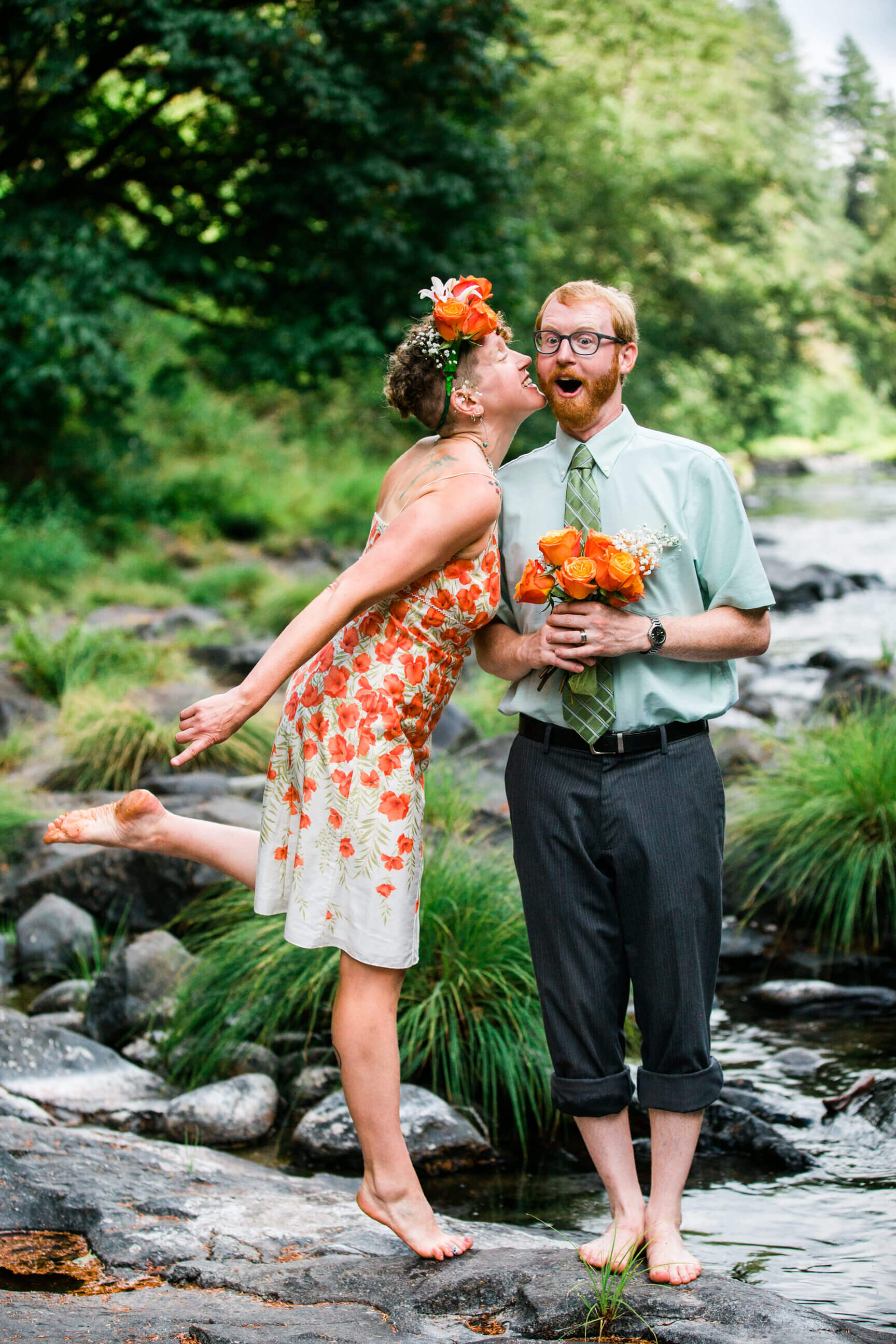 A groom holds a bouquet and is surprised as his bride leans in to kiss him on the Trask River near Tillamook Oregon