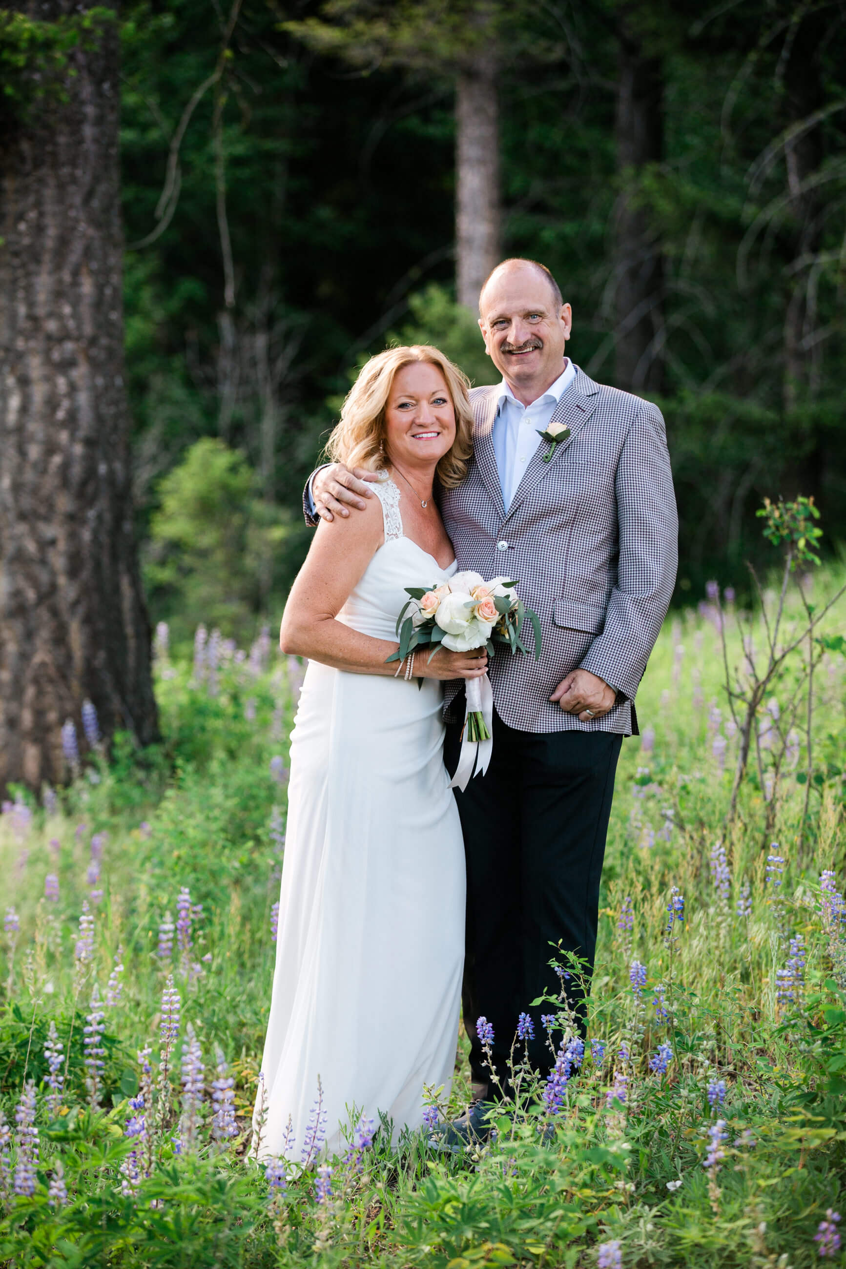 A bride and groom smile and stand in a field of lupine wildflowers during their intimate wedding at Triple Creek Ranch in Darby Montana