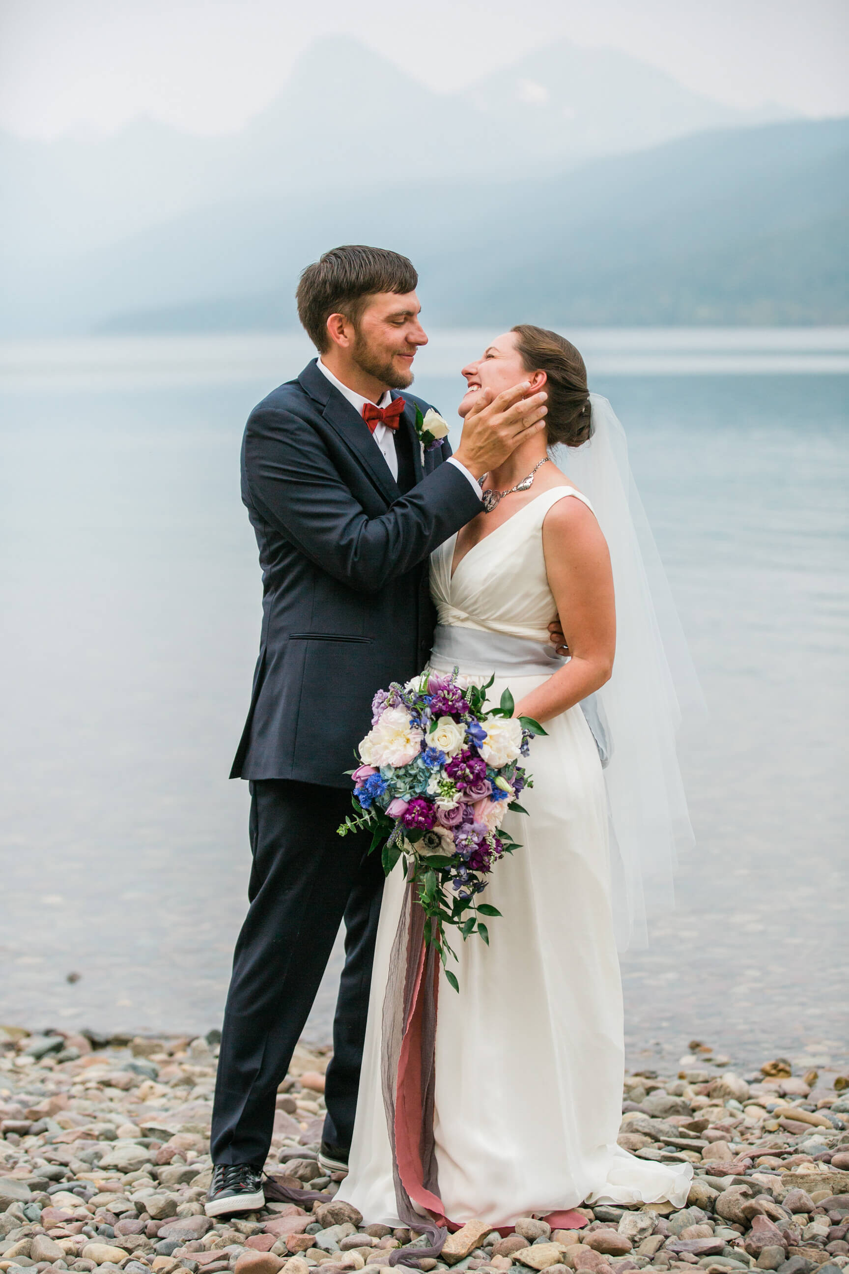 A groom tenderly holds the face of his bride as they stand on a rocky beach on Lake McDonald during their Glacier National Park elopement