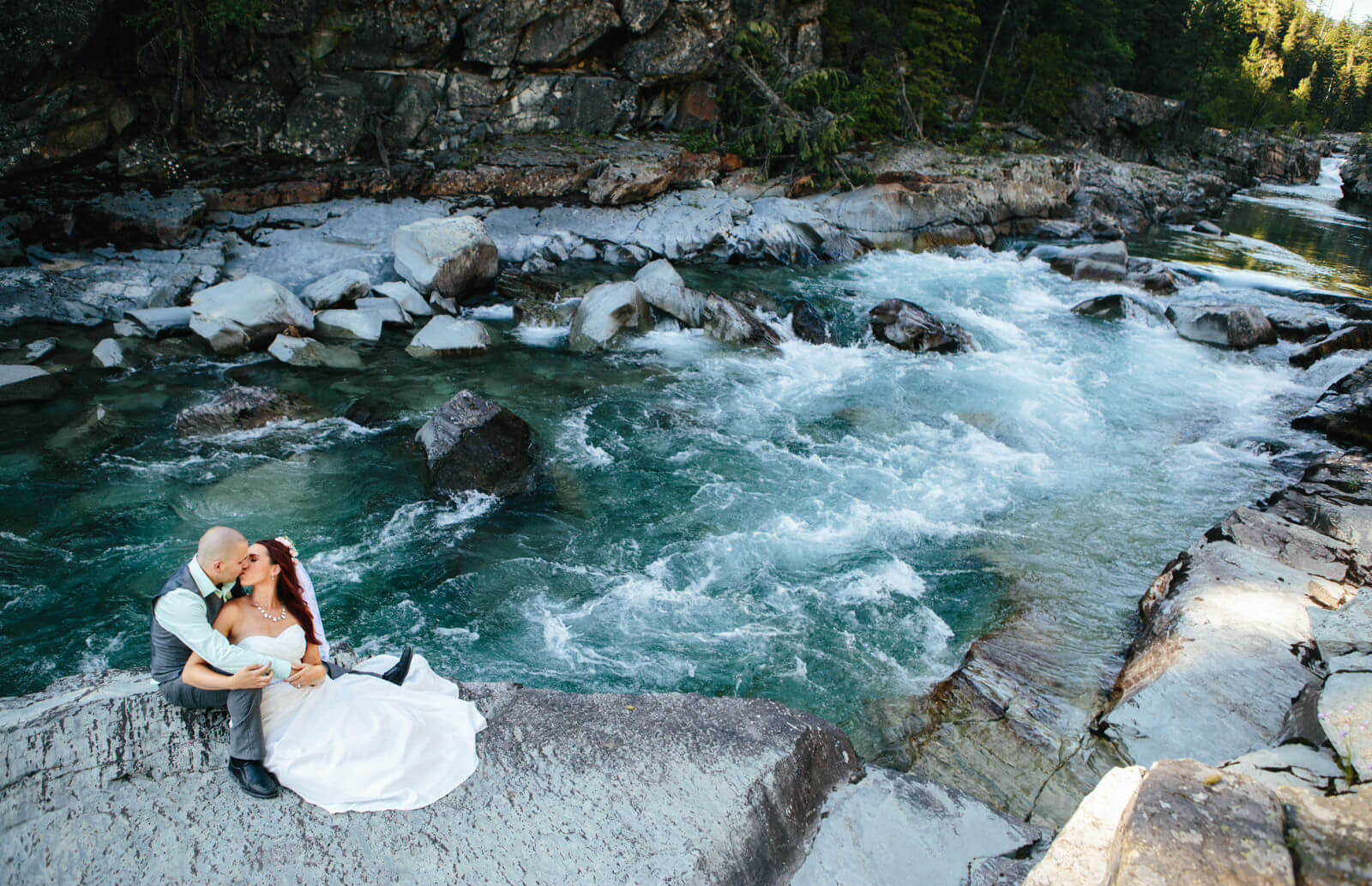 A bride and groom sit atop a giant boulder and kiss while a creek rages behind them at their Glacier National Park elopement in Montana